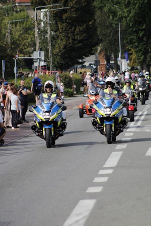 Widok dwóch policyjnych radiowozów na których jadą umundurowani policjanci po drodze a za nimi  jadą inne motocykle.