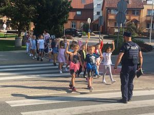 Zdjęcie przedstawia policjanta na oznakowanym przejściu dla pieszych  z grupą dzieci.