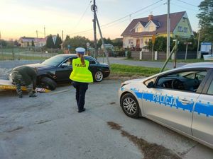 Zdjęcie przedstawia radiowóz w prawym roku oraz stojąca policjantkę w kamizelce jaskrawo zielonej a przed nią zielone auto z rozwalonym przednim kołem.