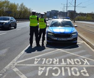 Zdjęcie przedstawia dwóch policjantów w umundurowaniu stojących na jezdni obok nich stojący oznakowany radiowóz a przed nim na jezdni napisany napis POLICJA KONTROLA