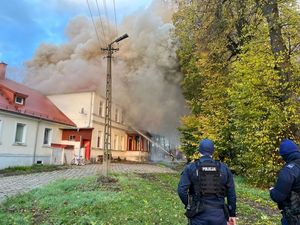 Zdjęcie przedstawia stojących tyłem dwóch umundurowanych policjantów oraz stojący przed nimi budynek cały w dymie.